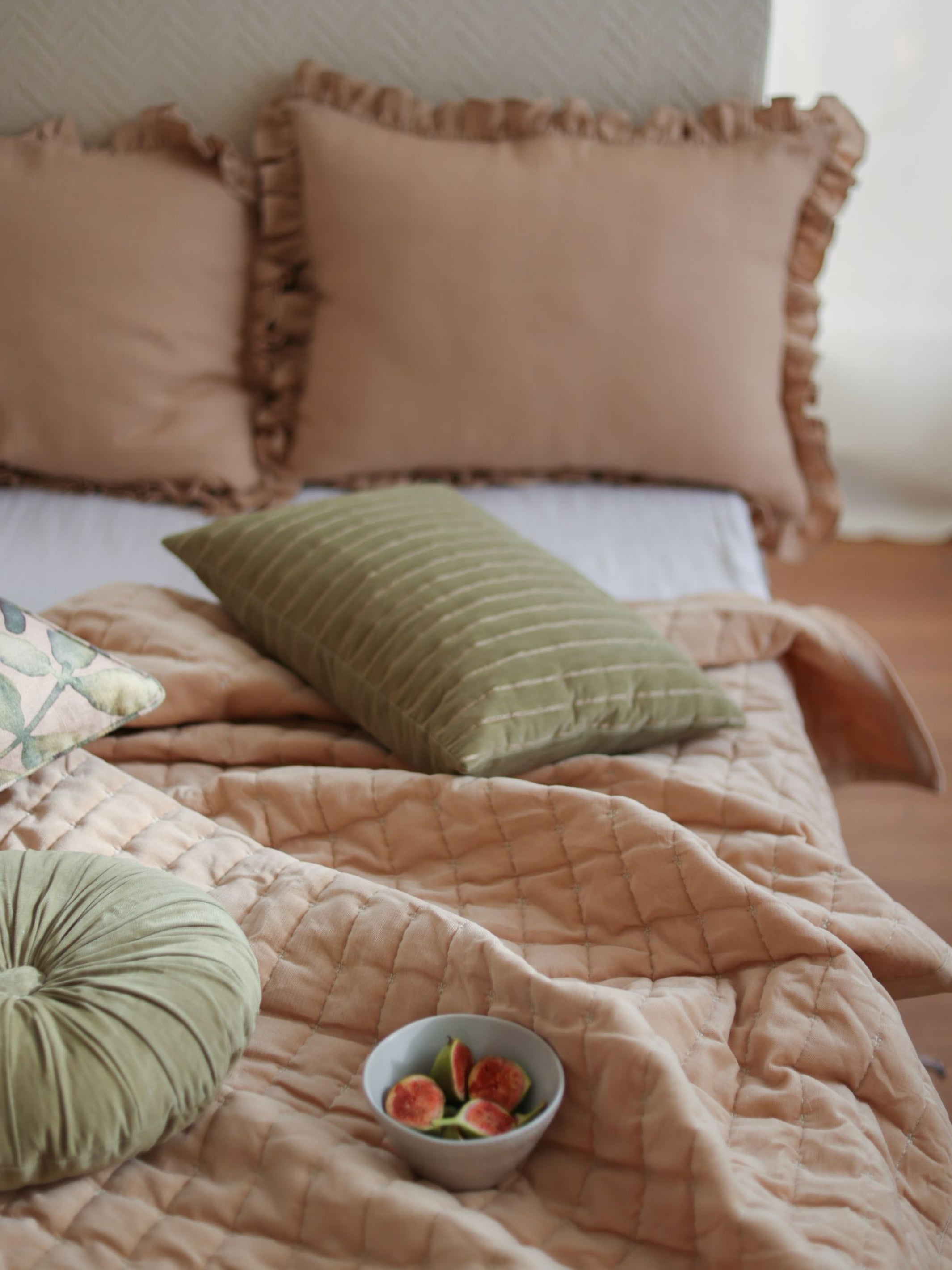 A cozy quilt with subtle star embroidery in a striped pattern is displayed on a bed, accompanied by two pillows and a bowl of fruit.