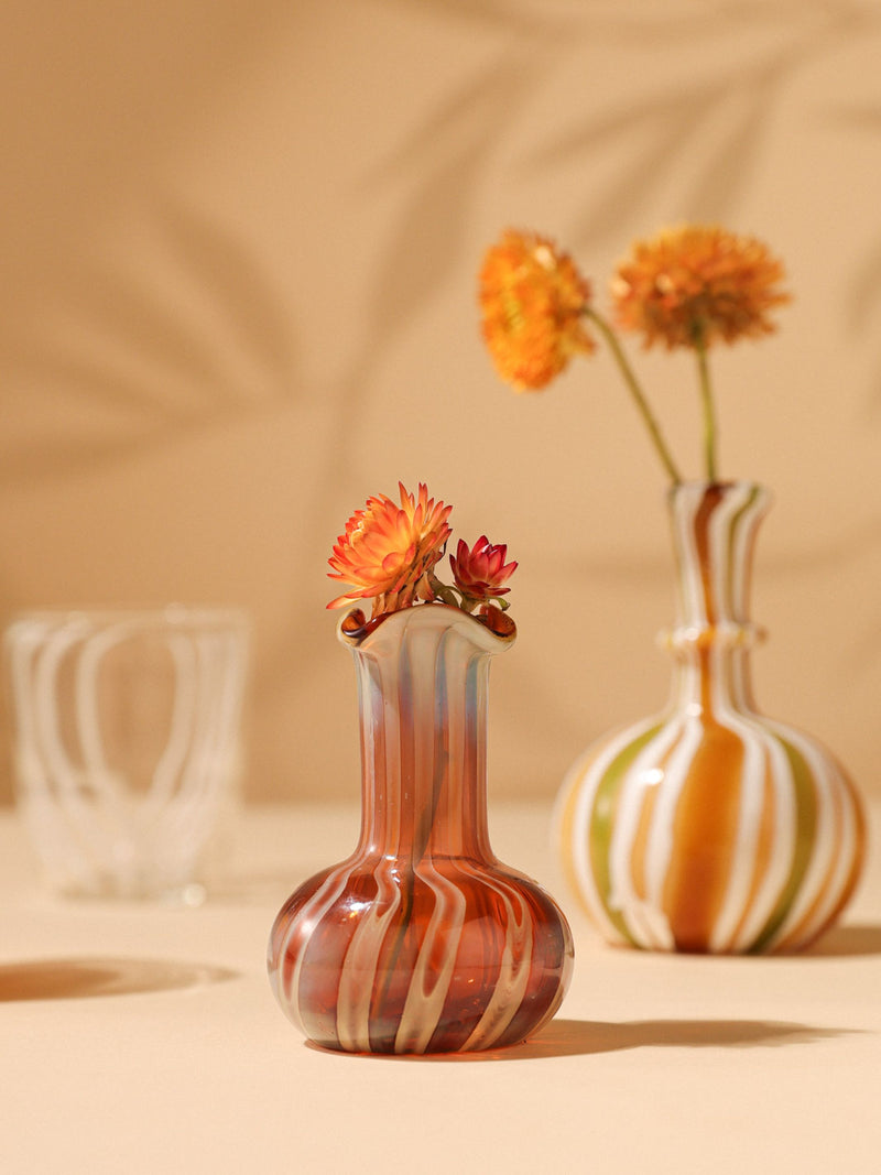 A handblown glass vase with earthy hues, featuring a swirling pattern, containing a bouquet of flowers. The vase is placed on a table with a blurred background.