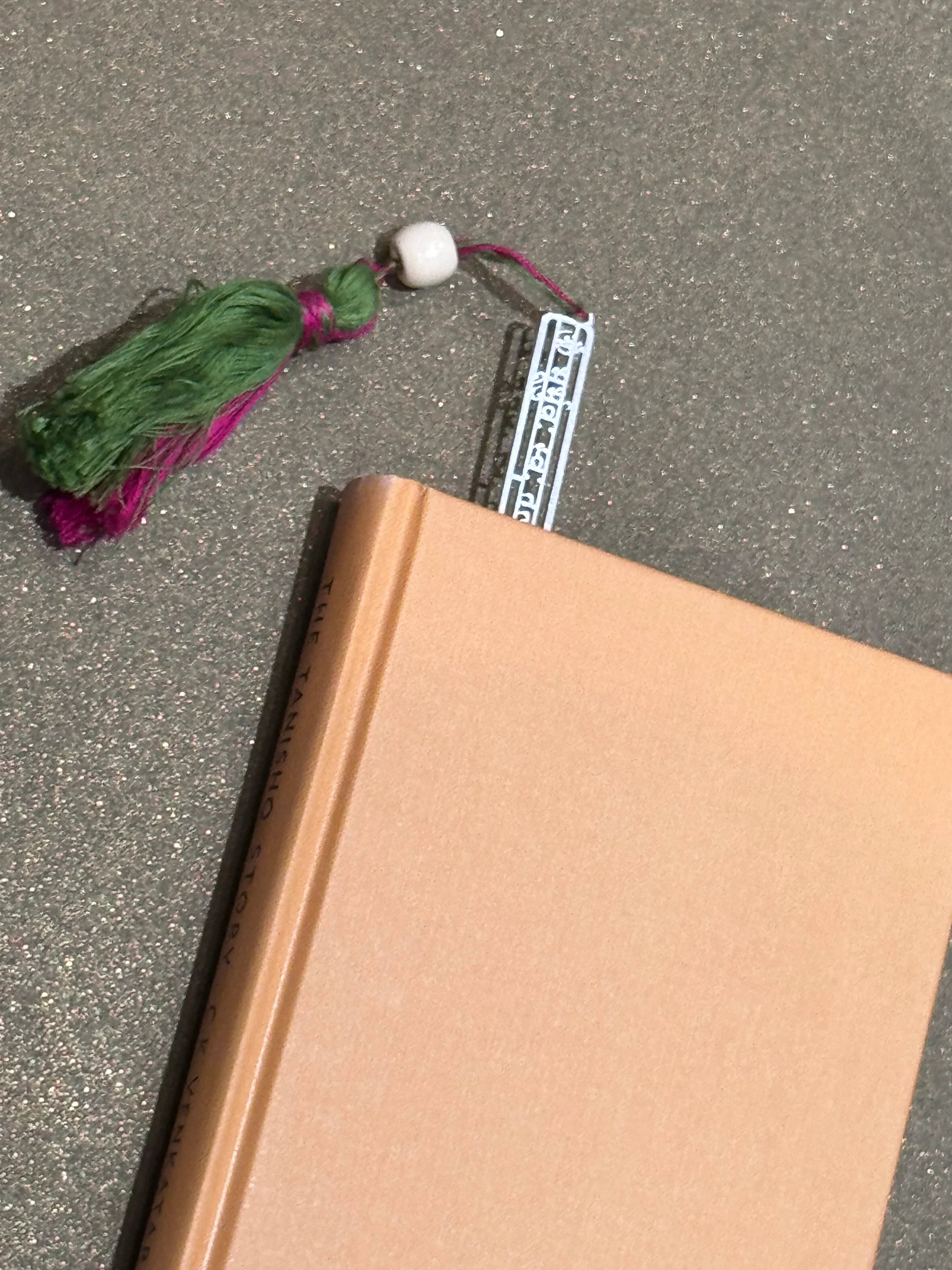Beige hardcover book with green and pink tassel bookmark on glitter surface, Sabera style