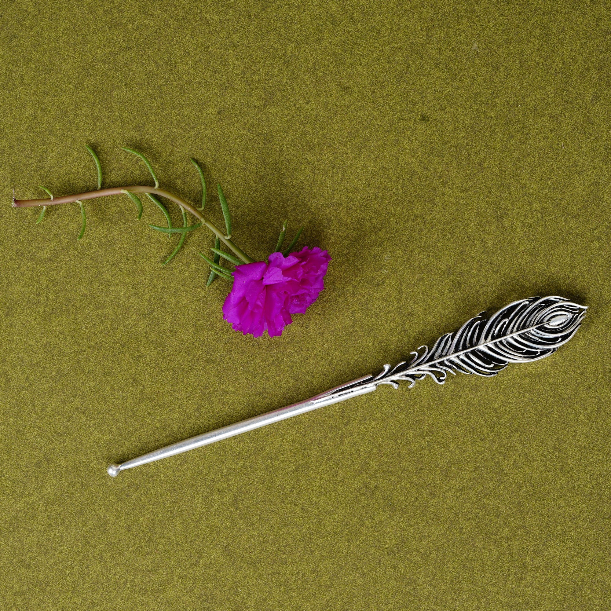 Metal peacock feather tilak stick and pink flower on olive green background, Sabera product