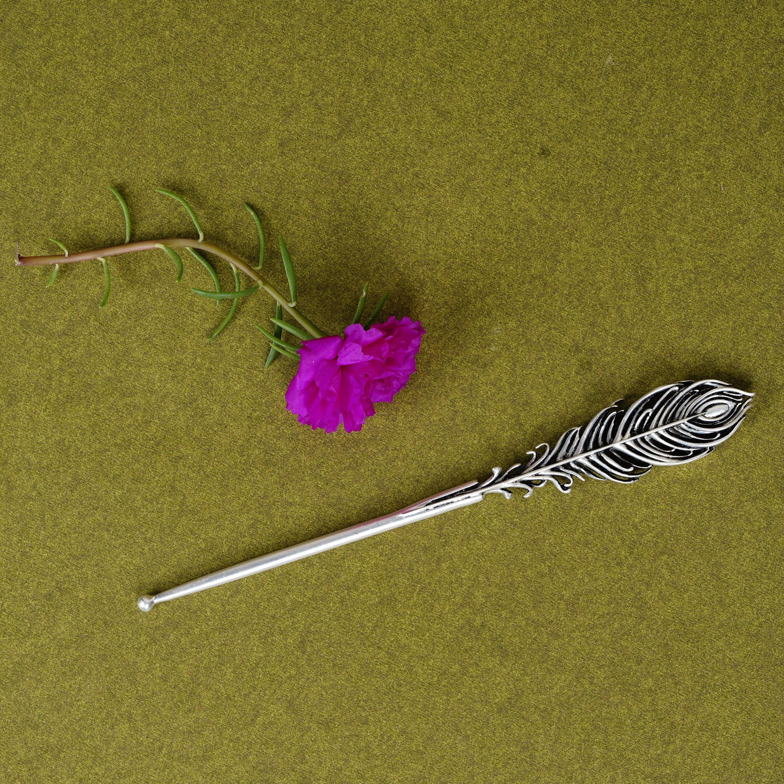 Metal peacock feather tilak stick and pink flower on olive green background, Sabera product