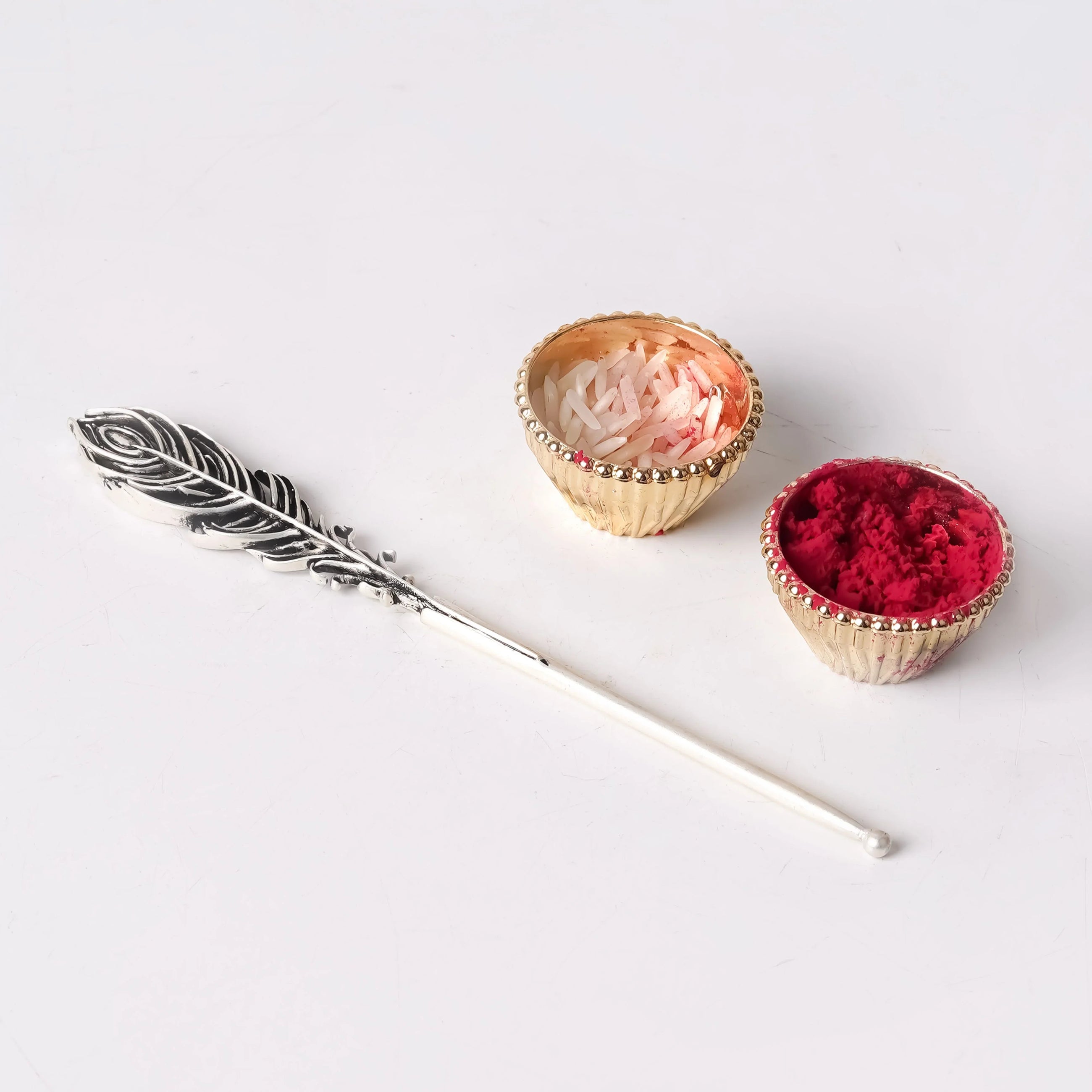 Peacock feather tilak stick with brass bowls of rice and red kumkum on white background