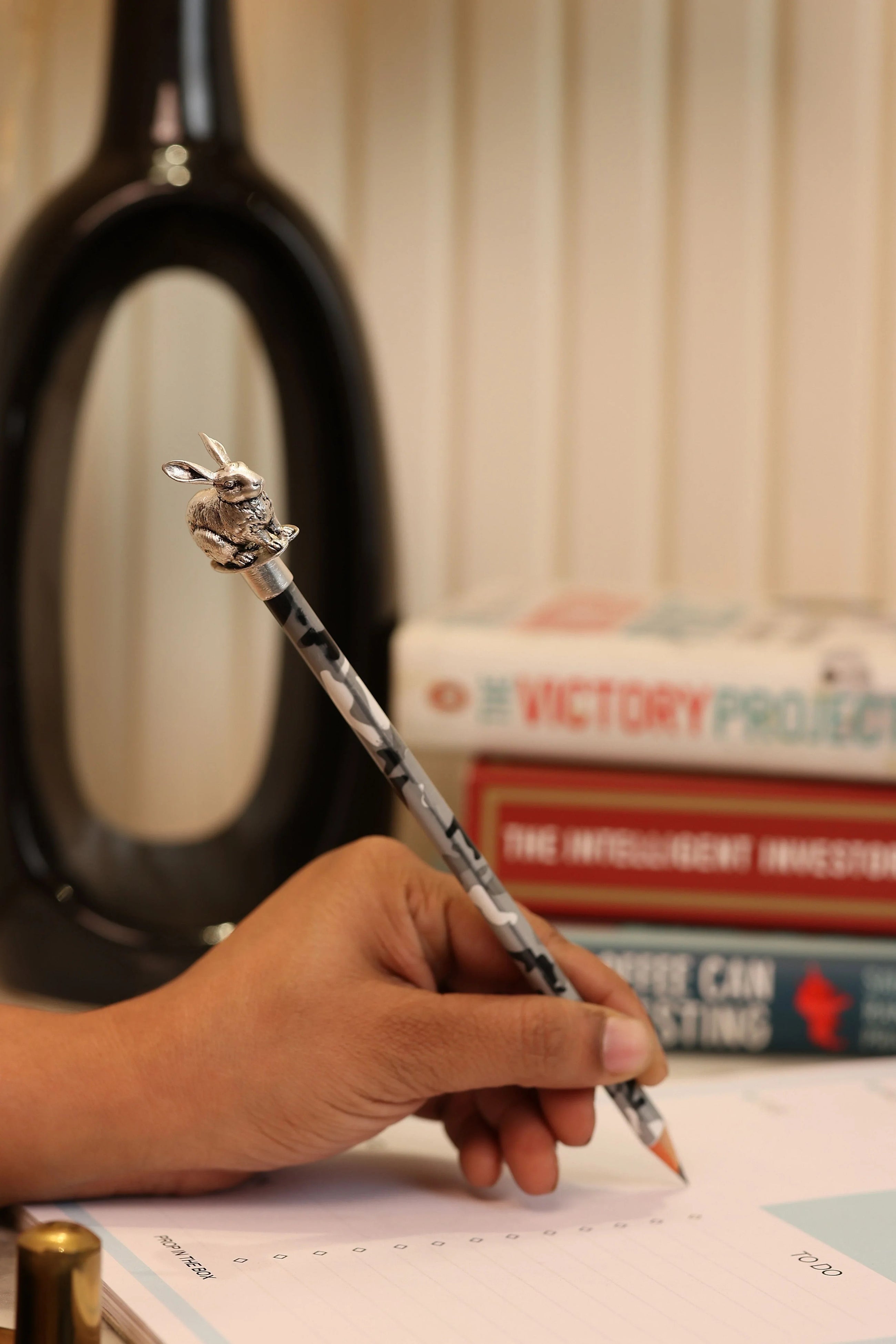 Hand holding pencil with Sabera silver bunny topper, books and notepad in background