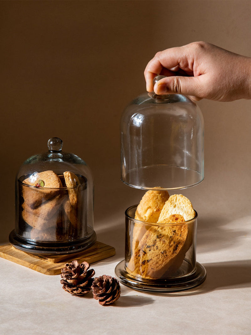 Hand placing a glass cloche over a container of cookies on a wooden board with pine cones.